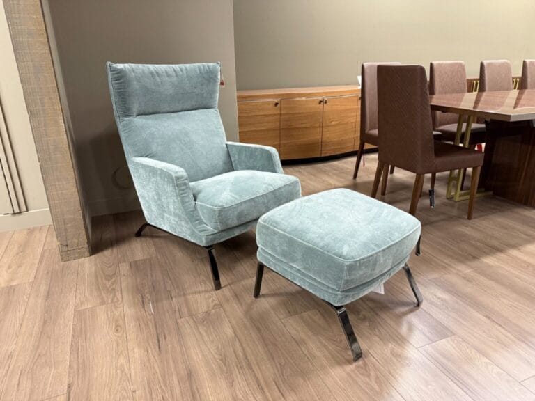 A light blue upholstered armchair with matching ottoman on metal legs, positioned on a wooden floor near a wooden cabinet and a dining table with brown chairs.