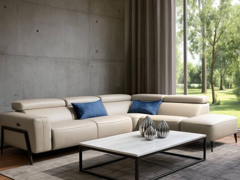 A modern living room with a beige sectional sofa, blue accent pillows, a white coffee table with metallic decor, and large windows overlooking green trees.