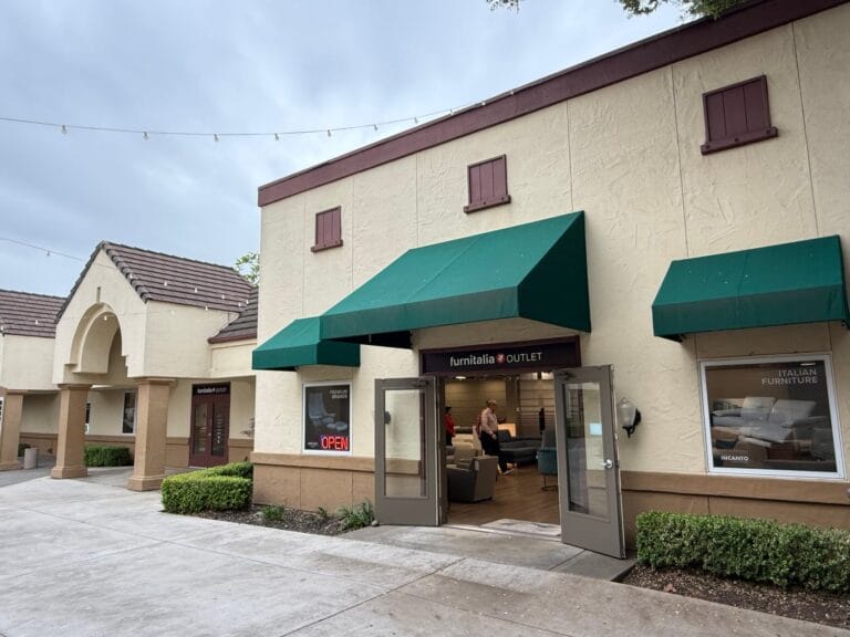 A furniture store with green awnings and open double doors, displaying sofas inside. Sidewalk and trimmed bushes are in front of the building.