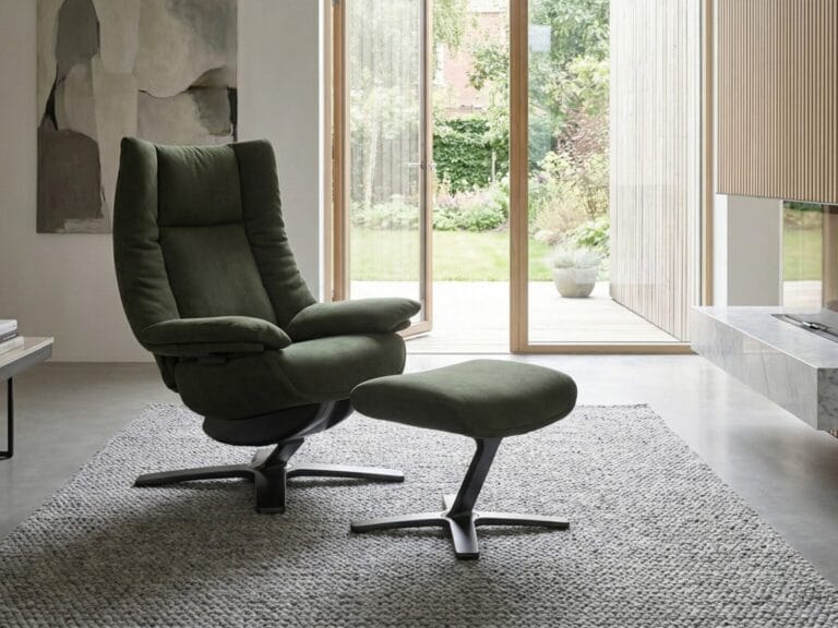 A green recliner chair with matching ottoman sits on a textured gray rug in a modern living room with large glass doors overlooking a garden.