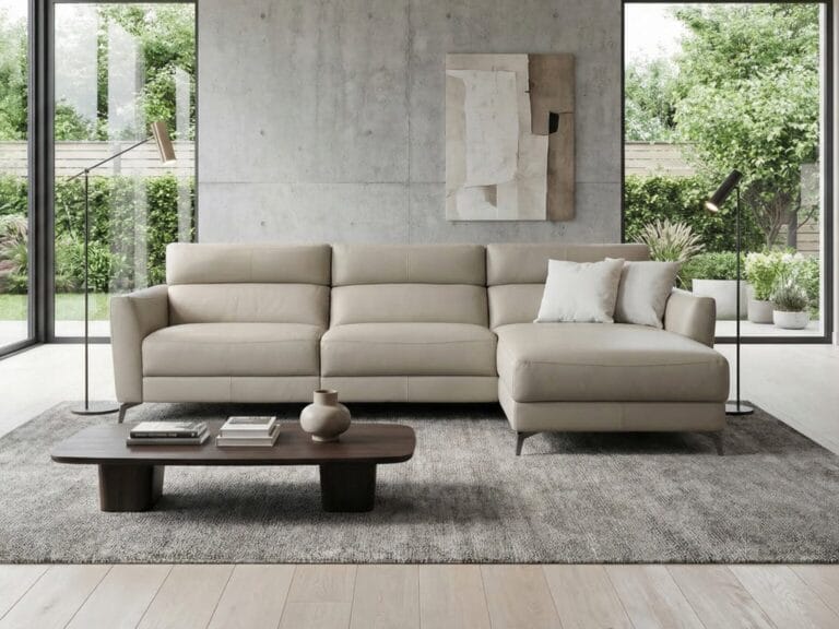 Modern living room with a beige sectional sofa, two white pillows, a low wooden coffee table with books, and large windows showing a garden view.