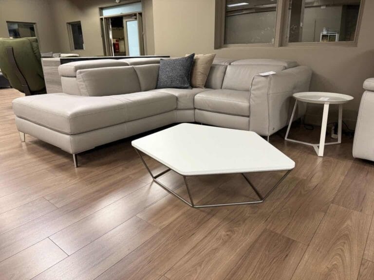 A modern living room with a light gray sectional sofa, assorted throw pillows, a hexagonal white coffee table, and a small round side table on wood flooring.
