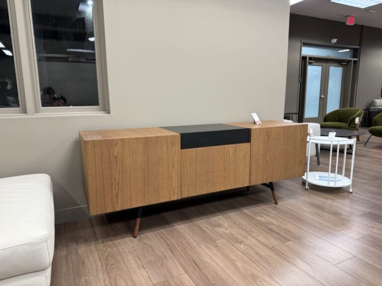 A wooden sideboard with angled legs and a black center panel is displayed in a showroom with wood flooring and neutral-colored walls.
