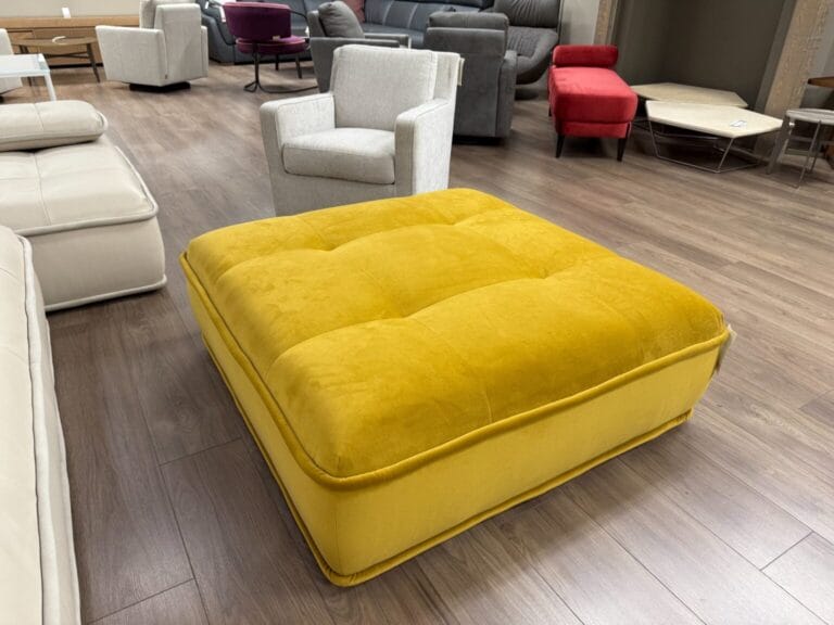 A large yellow square ottoman is placed on a wooden floor in a furniture showroom with various sofas and chairs in the background.
