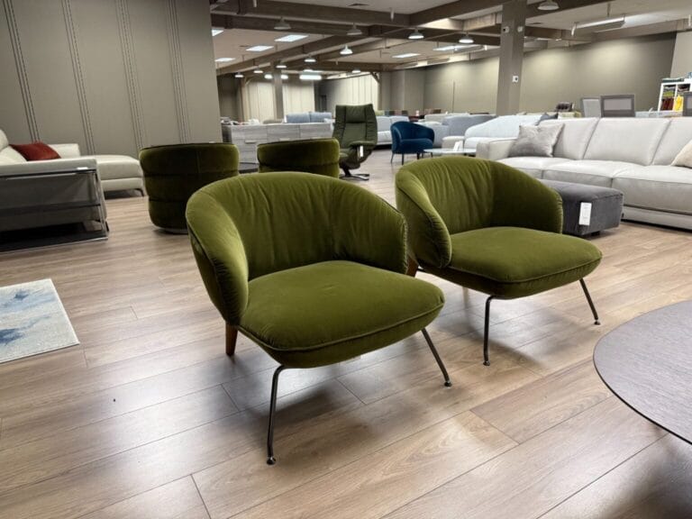 Two green velvet armchairs with black legs are placed on a wood floor in a modern furniture showroom with sofas and chairs in the background.