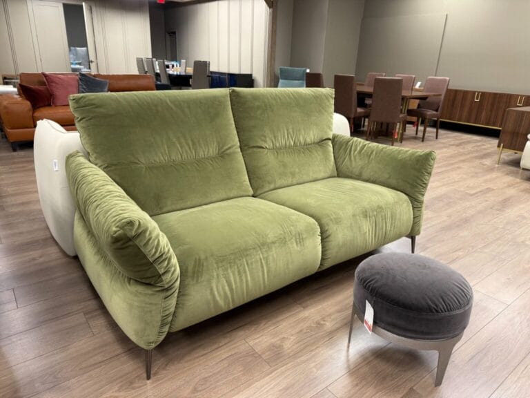 A green upholstered loveseat with metal legs is positioned near a round gray ottoman in a modern furniture showroom with wood flooring.