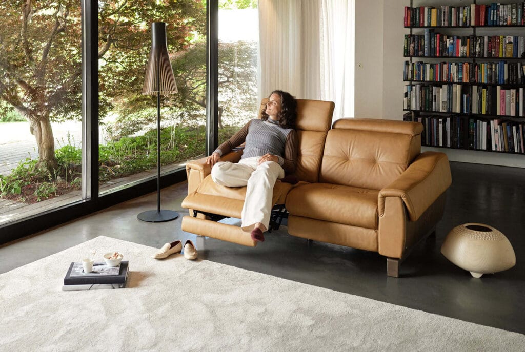 A person relaxes on a tan leather recliner in a modern living room with large windows, a bookshelf, and a floor lamp.