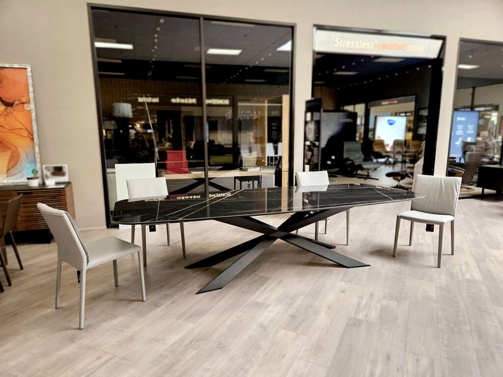 Modern black marble dining table with a geometric metal base, surrounded by four white chairs, displayed in a showroom with large glass windows and wood flooring.
