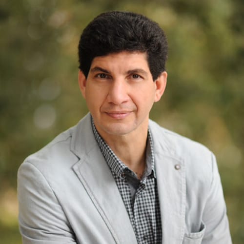 A man with short dark hair wearing a gray blazer over a checked shirt, seated outdoors with a blurred green background.