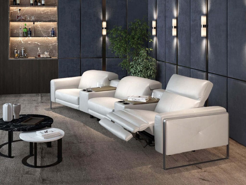 A modern living room with three white leather recliner chairs, two small round black tables, a bar shelf with bottles, and a plant against dark paneled walls.