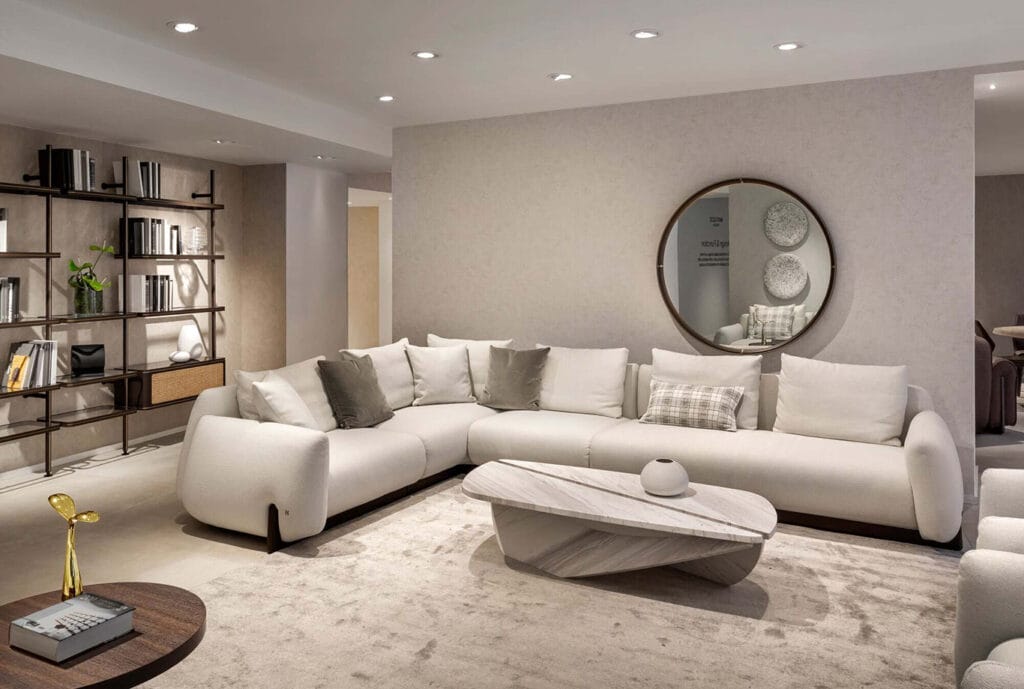 Modern living room with a large white sectional sofa, neutral cushions, round marble coffee table, open bookshelf, and a round wall mirror above the sofa.