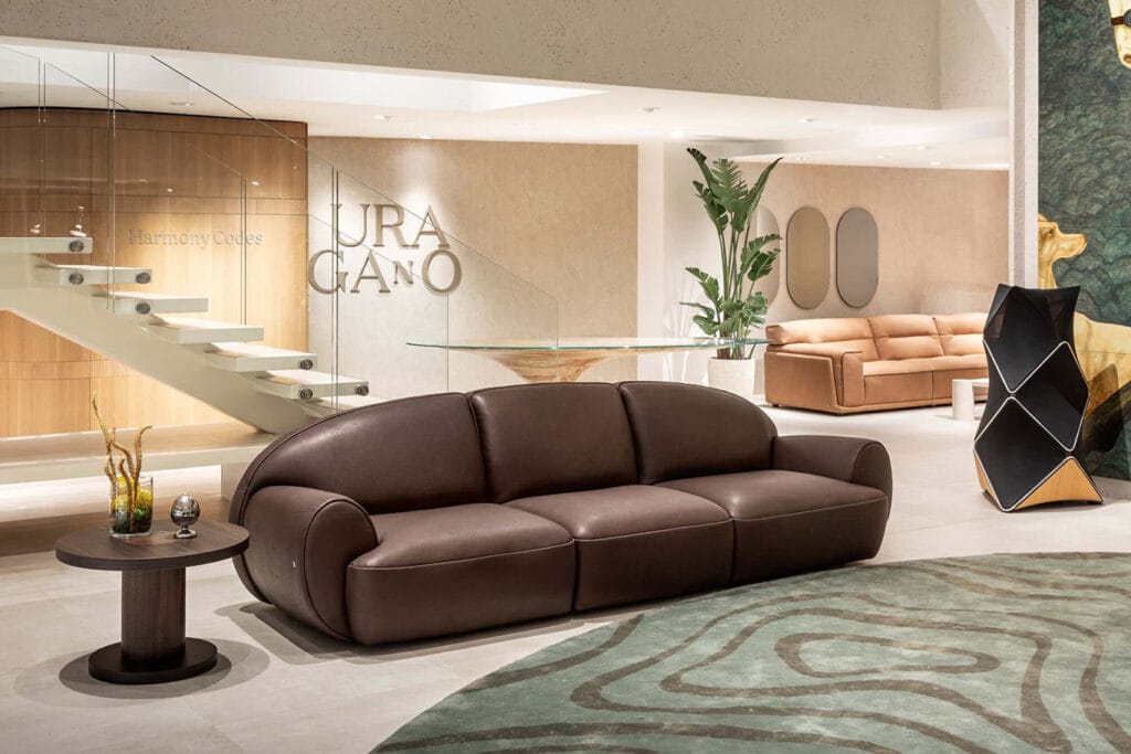 Modern living room with a brown leather sofa, round side table, indoor plants, and beige décor; glass staircase and artwork are visible in the background.