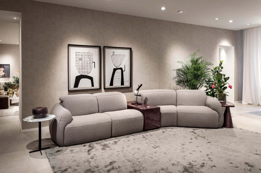Modern living room with a light gray sectional sofa, abstract art on the wall, side tables, a table lamp, and several potted plants on a large rug.