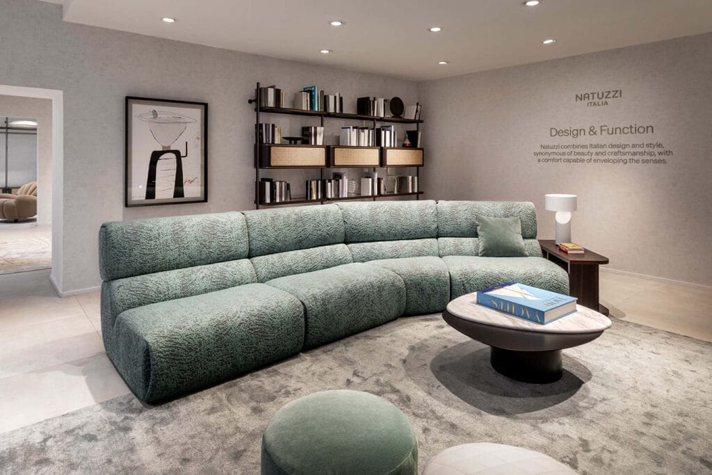 Modern living room with a large green sectional sofa, round coffee table with books, shelving unit with decor, and abstract wall art, under recessed ceiling lights.