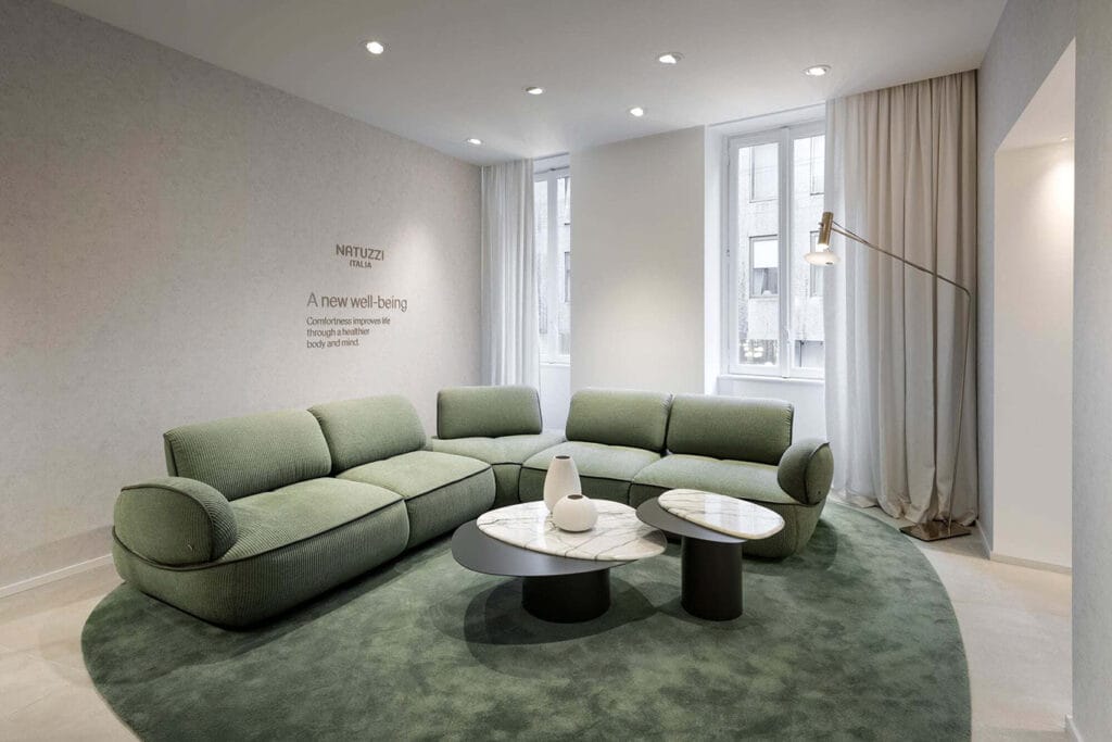 Modern living room with green sectional sofa, two round coffee tables with a white vase, floor lamp, large windows, and light curtains on a circular green rug.