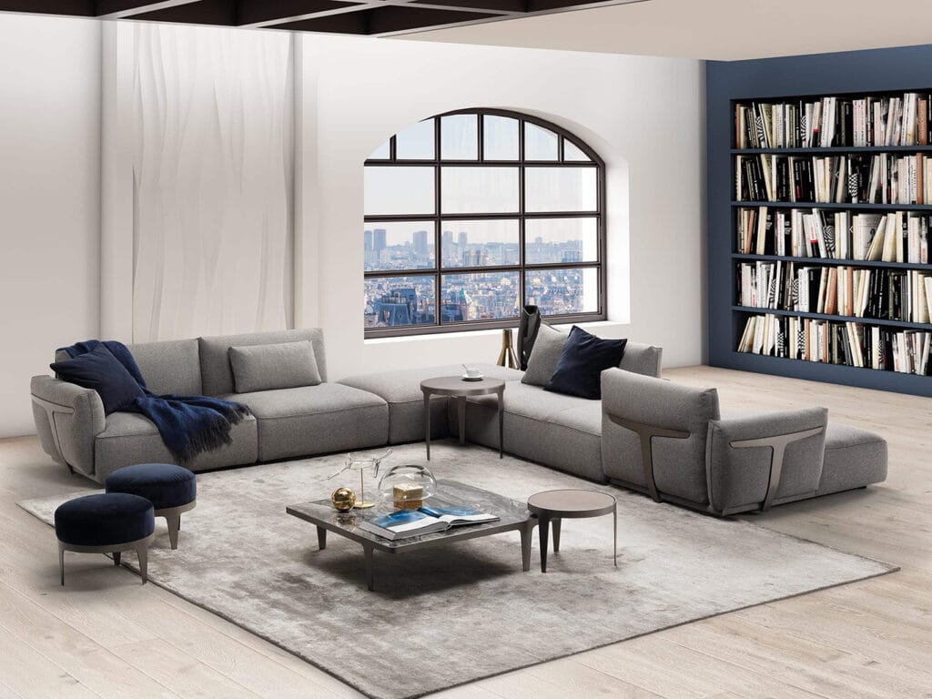 Modern living room with a grey sectional sofa, armchair, coffee table, ottomans, bookshelves, and large arched window overlooking a cityscape.