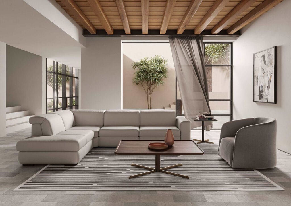 Modern living room with a white sectional sofa, gray armchair, wooden coffee table, striped rug, large windows, potted plant, and wall art under a wooden ceiling.