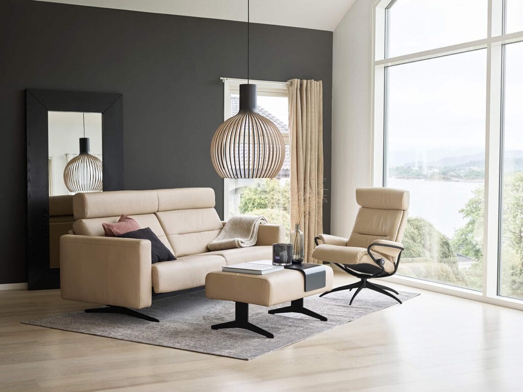 Modern living room with beige sofa and armchair, ottoman, large window with view, black accent wall, floor mirror, and pendant light.