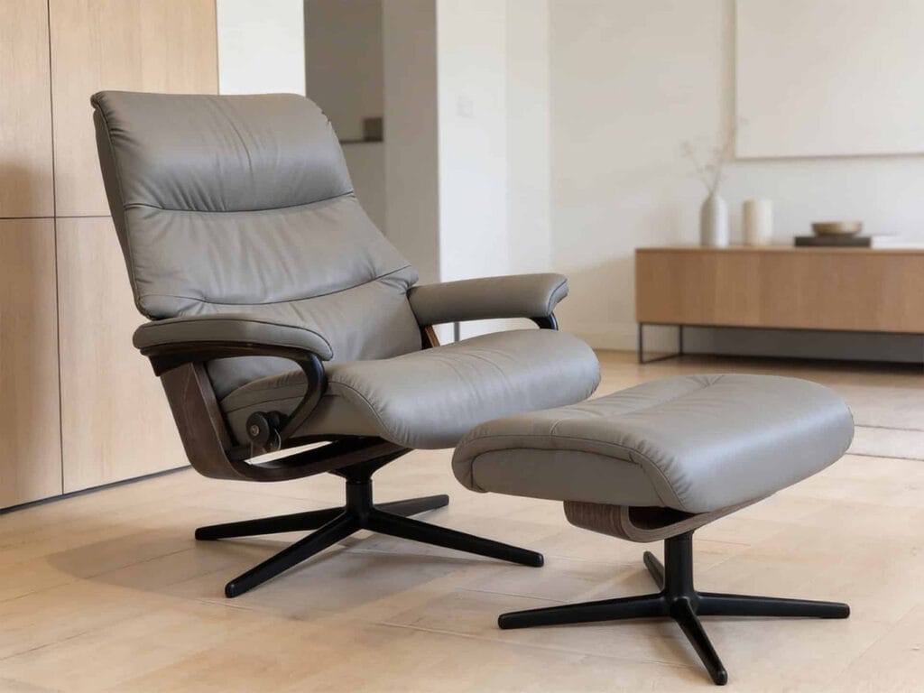 A modern gray leather recliner chair with matching ottoman, both on black swivel bases, placed in a minimalist living room with light wood flooring.
