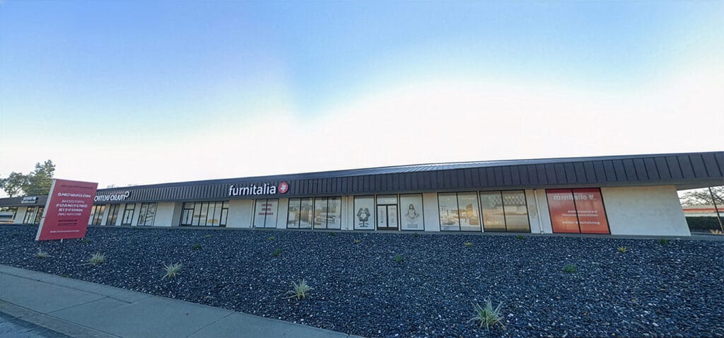 A single-story building with "furnitalia" signage, large windows, and a landscaped rock garden in front. A red sign stands near the sidewalk.